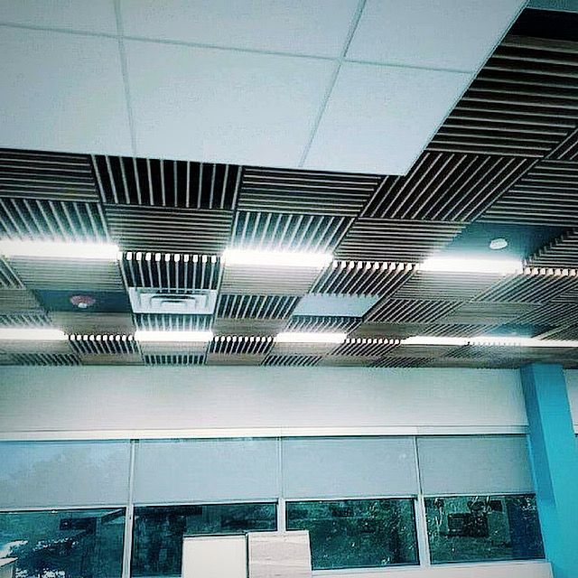 A ceiling with alternating wood slat and white square panels, with recessed lighting, above a windowed wall.