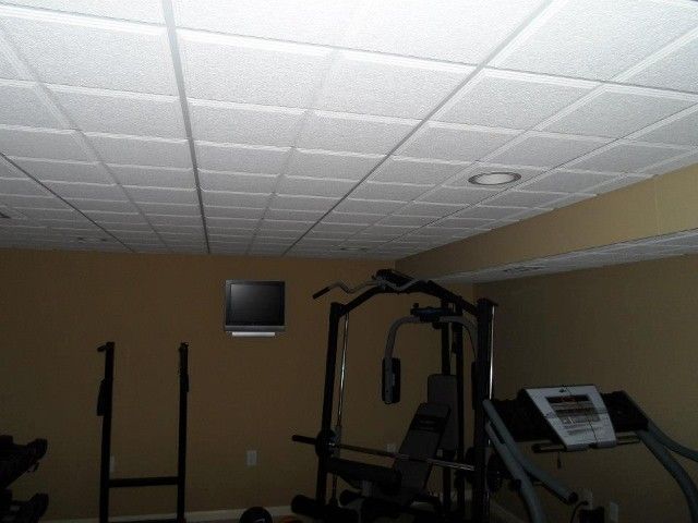 Home gym with a weight machine and treadmill under a tile ceiling. A small TV is mounted on the wall.