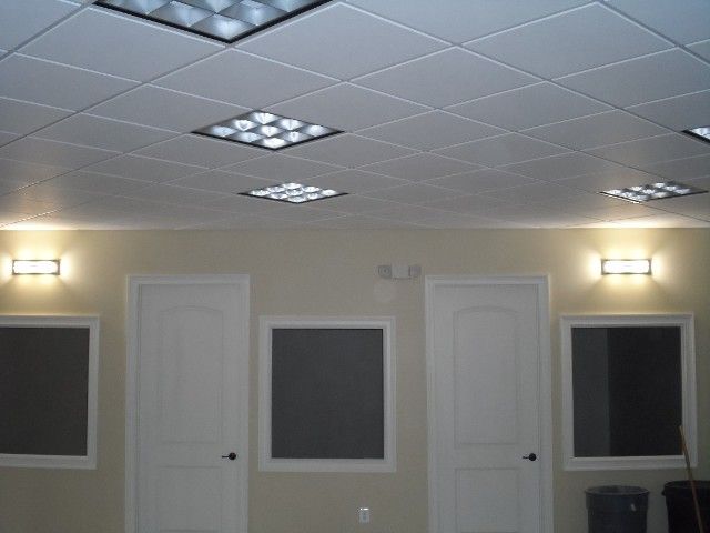 A room with a dropped ceiling, fluorescent lights, doors, windows, and wall sconces.