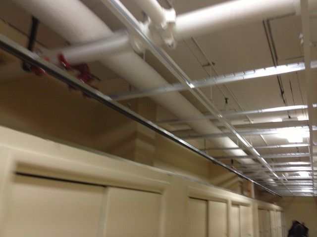Hallway ceiling with pipes, metal supports, and recessed lighting. Cream walls and closed doors.