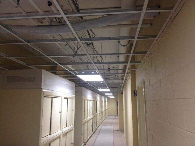 Hallway with exposed ceiling grid, fluorescent lights, lockers, and tan walls.