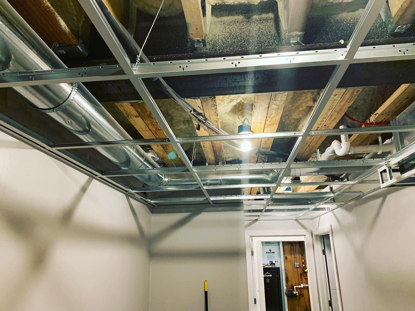 Metal grid ceiling framework with exposed ductwork, plumbing, and wood beams. A lightbulb hangs.