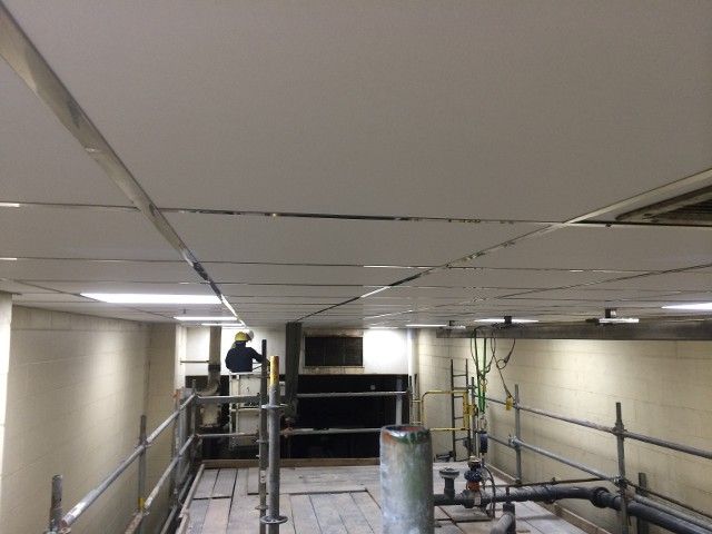 Person working on a dropped ceiling in a construction area, scaffolding, metal framework, and utility pipes visible.