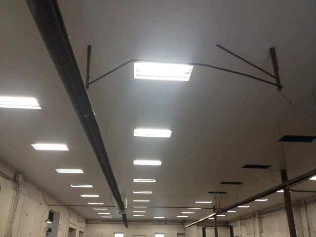 Interior of a building with a white ceiling. Rows of square, bright lights are installed. Black metal beams hang down.
