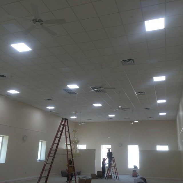 Interior view of a room with ceiling lights, ladders, and people working. White walls and ceiling.