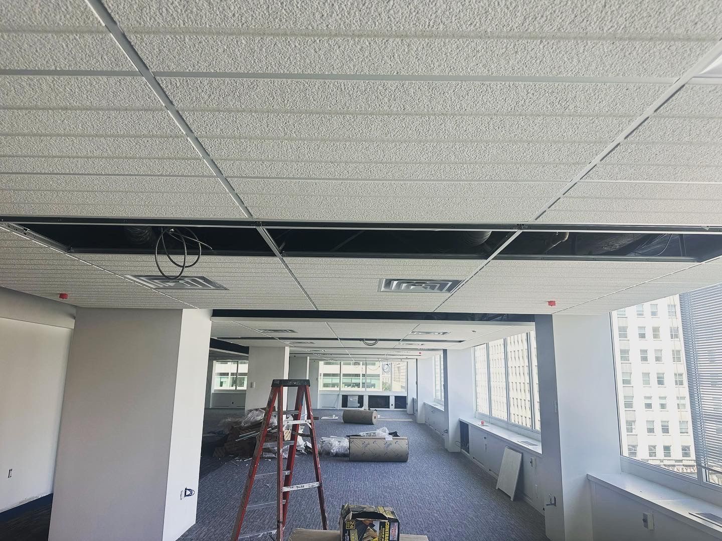 Office ceiling with missing tiles, exposing wiring, overlooking a city. Ladder visible.