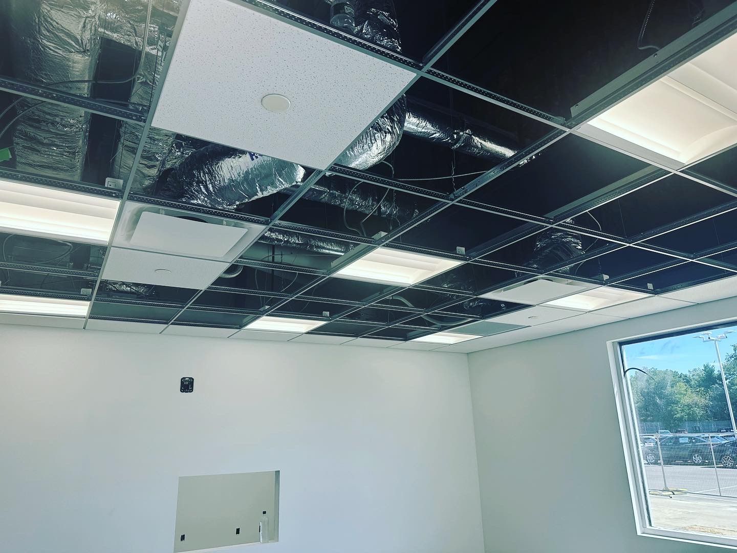 Interior view of a room under construction with a drop ceiling, lighting, and a window. Walls are white.
