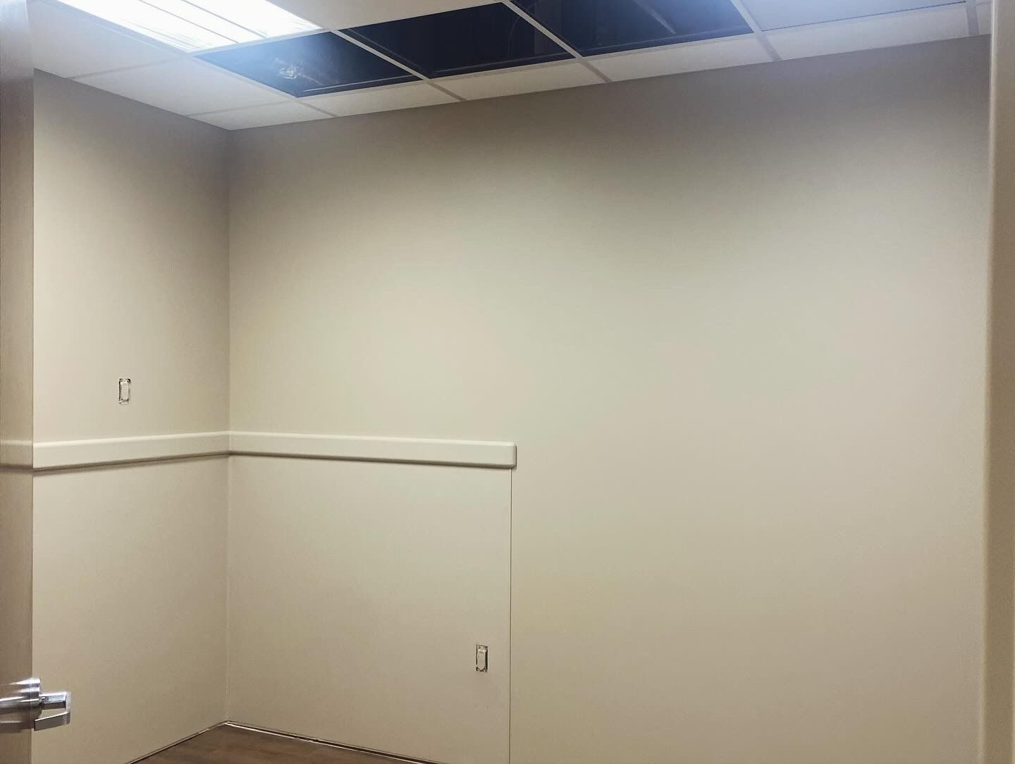 Empty, beige-painted room with a partial drop ceiling and recessed lighting. A door and a small opening are visible.