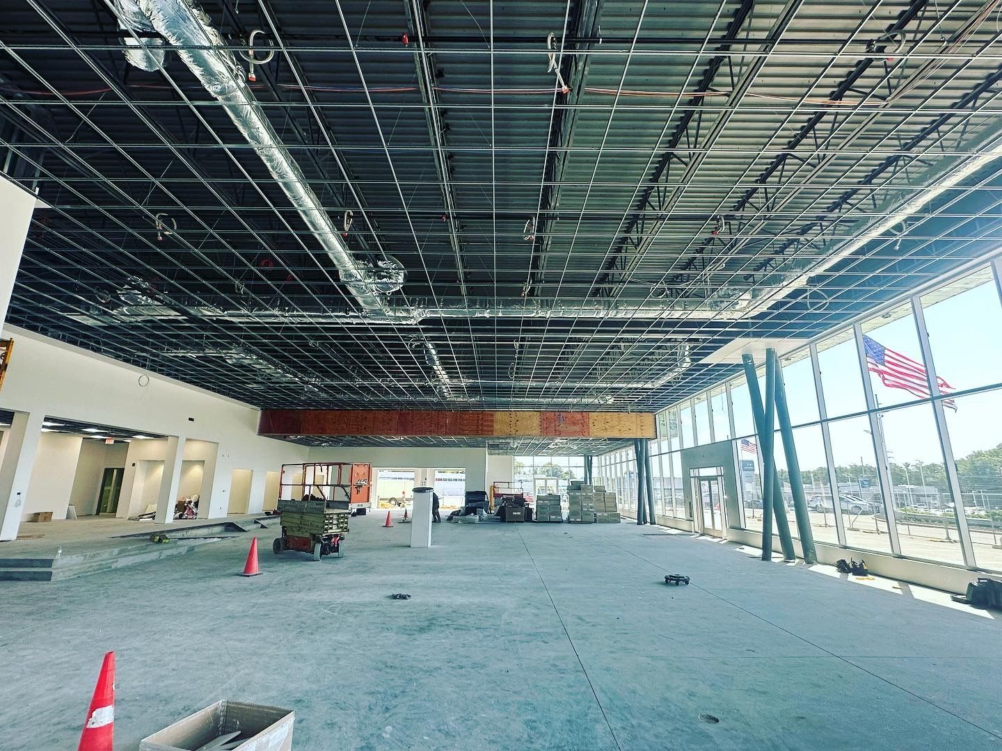 Interior of a building under construction, with exposed ceiling grid, large windows, and an American flag visible.