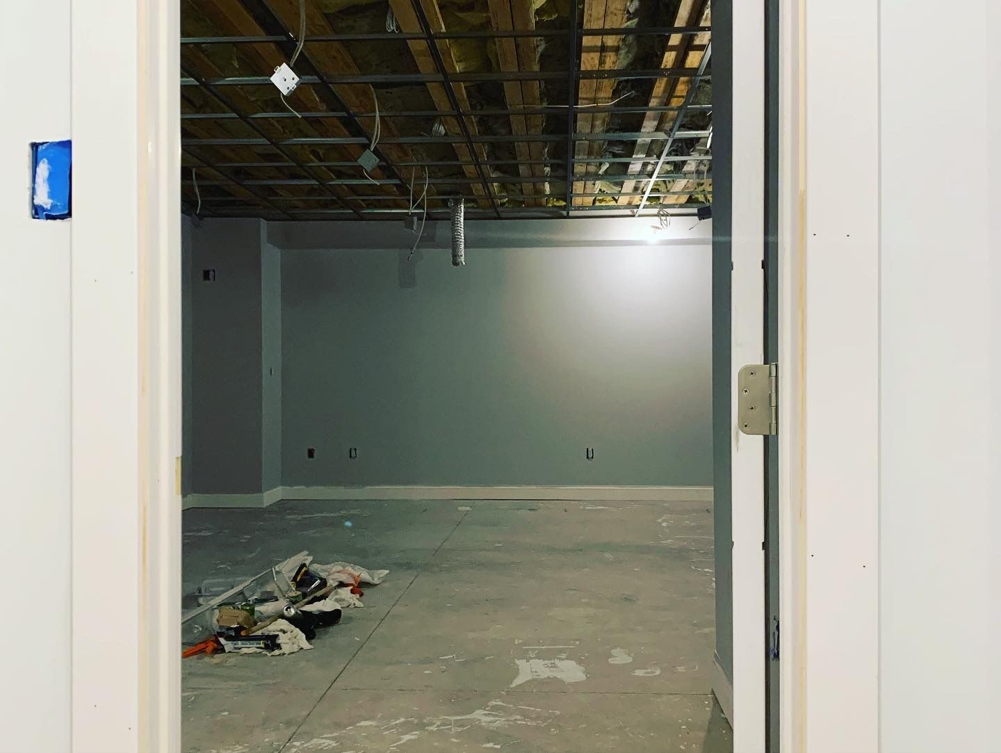 Room under construction; unfinished concrete floor, gray walls, open ceiling with exposed metal grid, and white door frame.