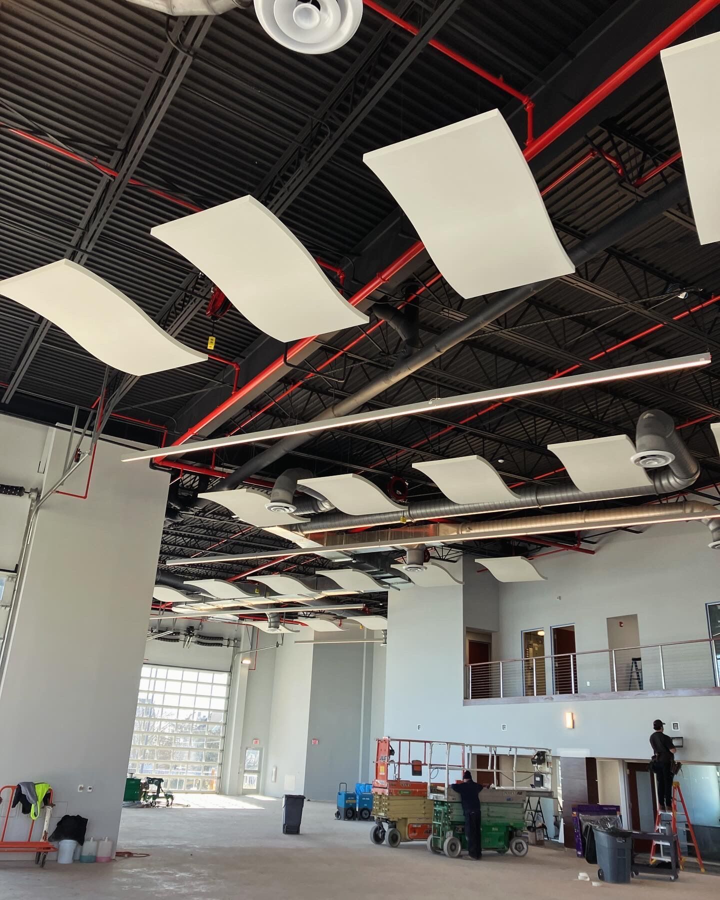 Interior of a building under construction, showing suspended acoustic panels, exposed pipes, and construction workers.