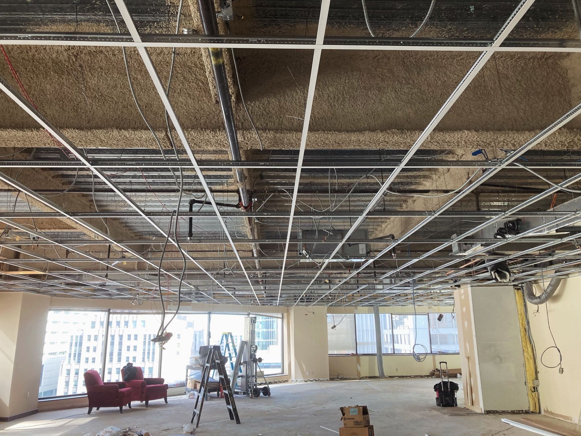 Construction site interior: ceiling grid, wiring, insulation, and windows. Empty room with red chairs.
