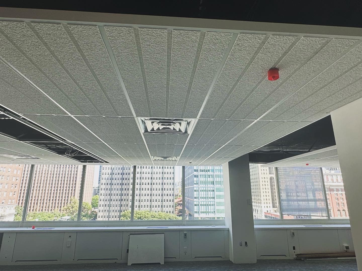 Office ceiling with white tiles, air vents, a fire alarm, and a window view of city buildings.