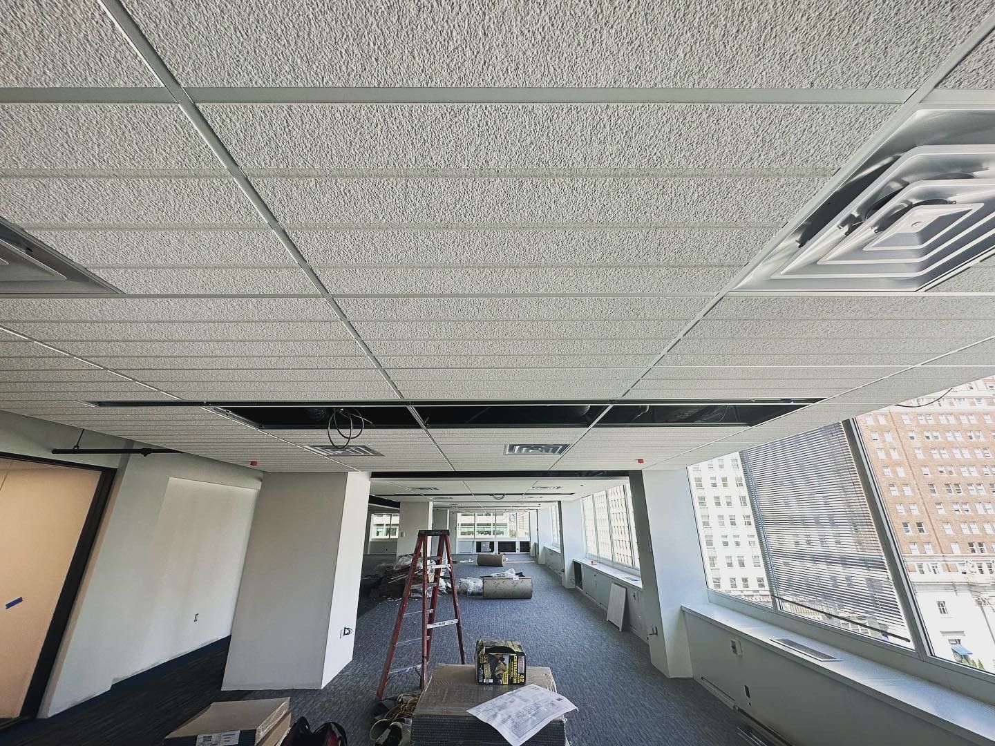 Office space ceiling with missing tiles; construction in progress. View toward windows.