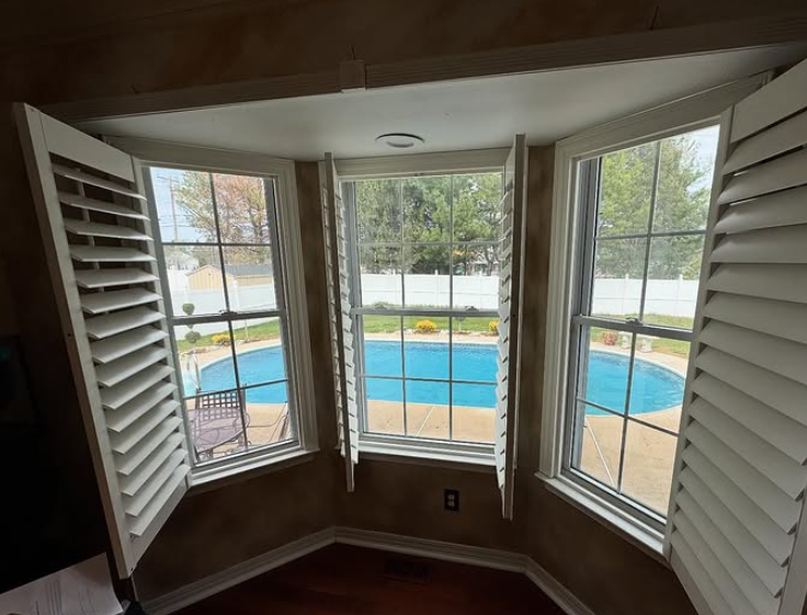 A room with white shuttered windows overlooking a blue pool and backyard.