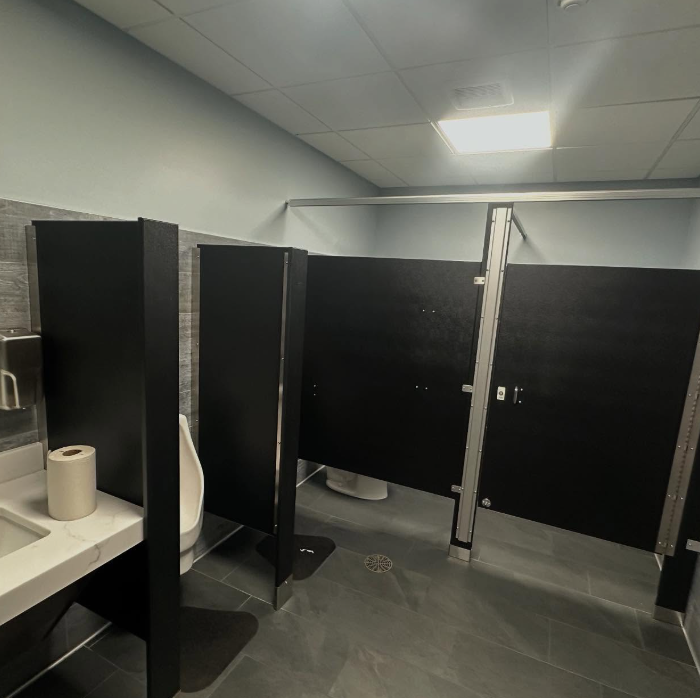 Public restroom with black stall doors and a urinal, gray tile floor, and a sink with a roll of toilet paper.