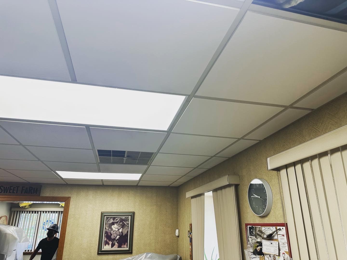 Office ceiling with recessed lighting and drop ceiling tiles. A wall with a clock, blinds, and a picture.