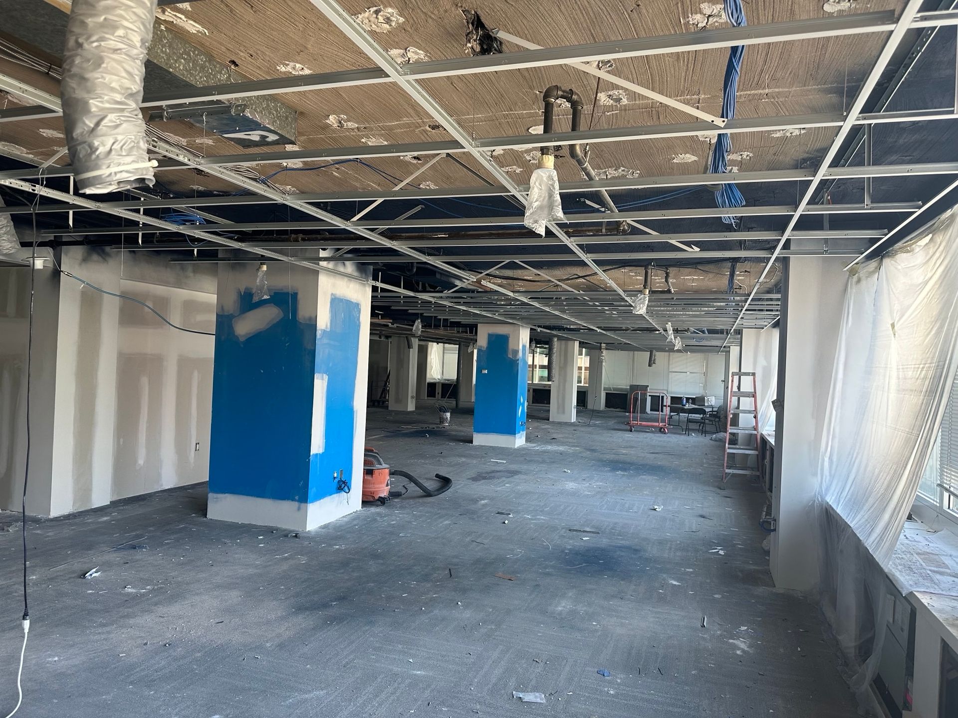 Interior space under construction; drywall, blue painted pillars, exposed ceiling grid.