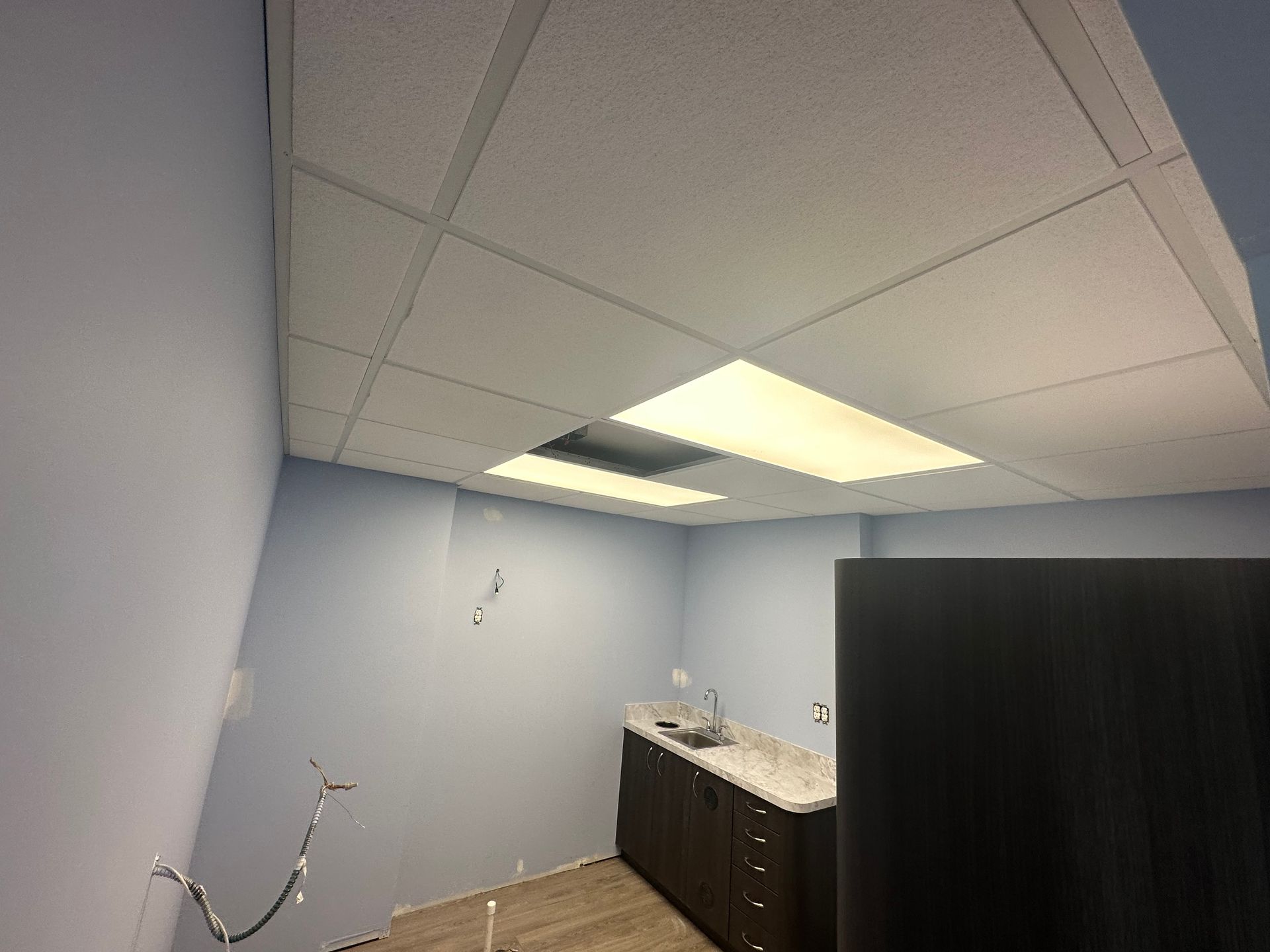 Room with drop ceiling, light fixtures, pale blue walls, dark cabinets, and wires.