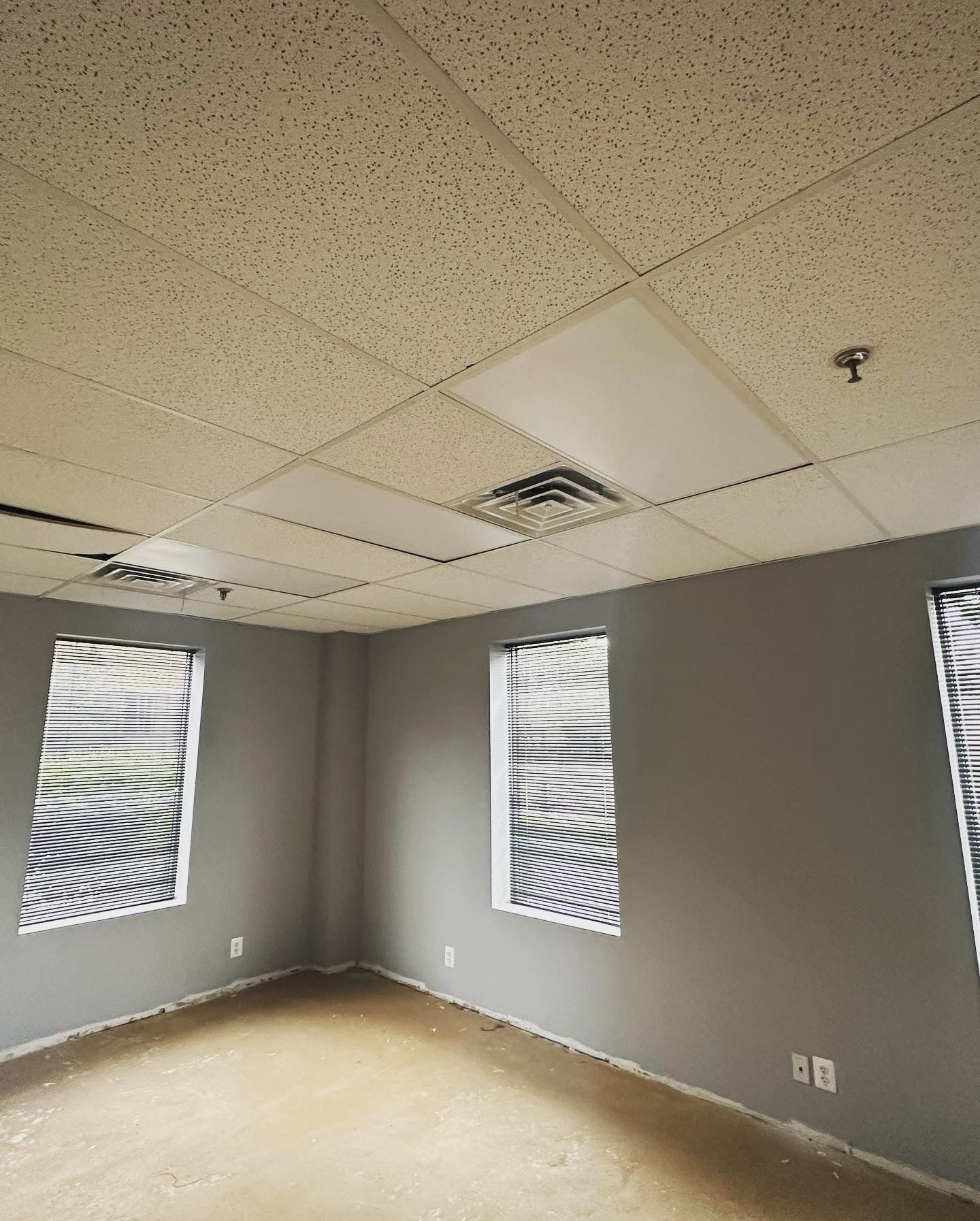 Empty office room with gray walls, three windows with blinds, and a drop ceiling.