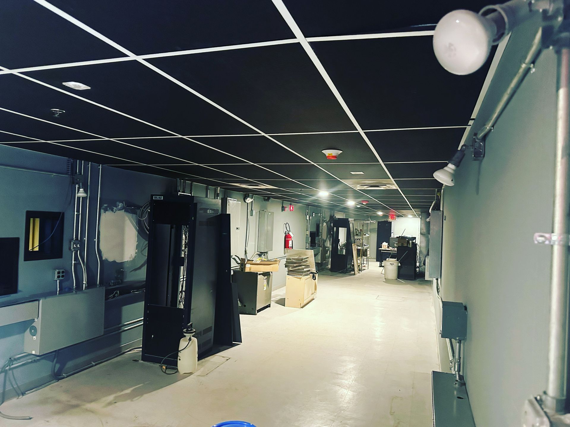 Long hallway with black ceiling grid, white walls, and industrial lighting; several doors and equipment.