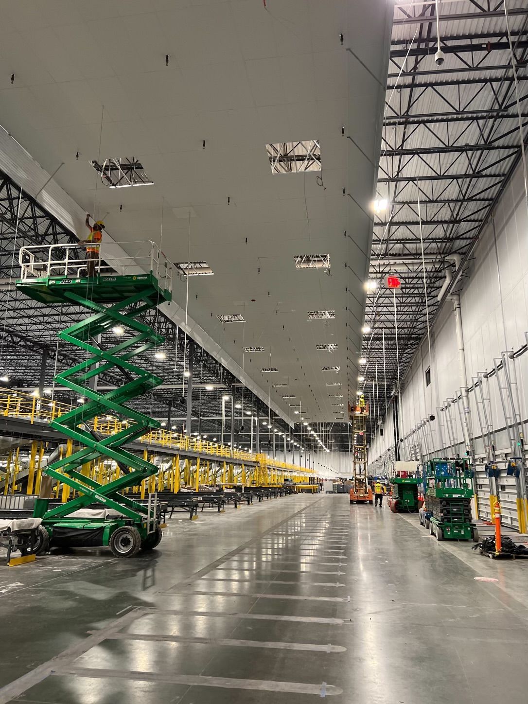 Warehouse interior with workers on lifts, installing ceiling panels. Green and yellow machinery, concrete floor.