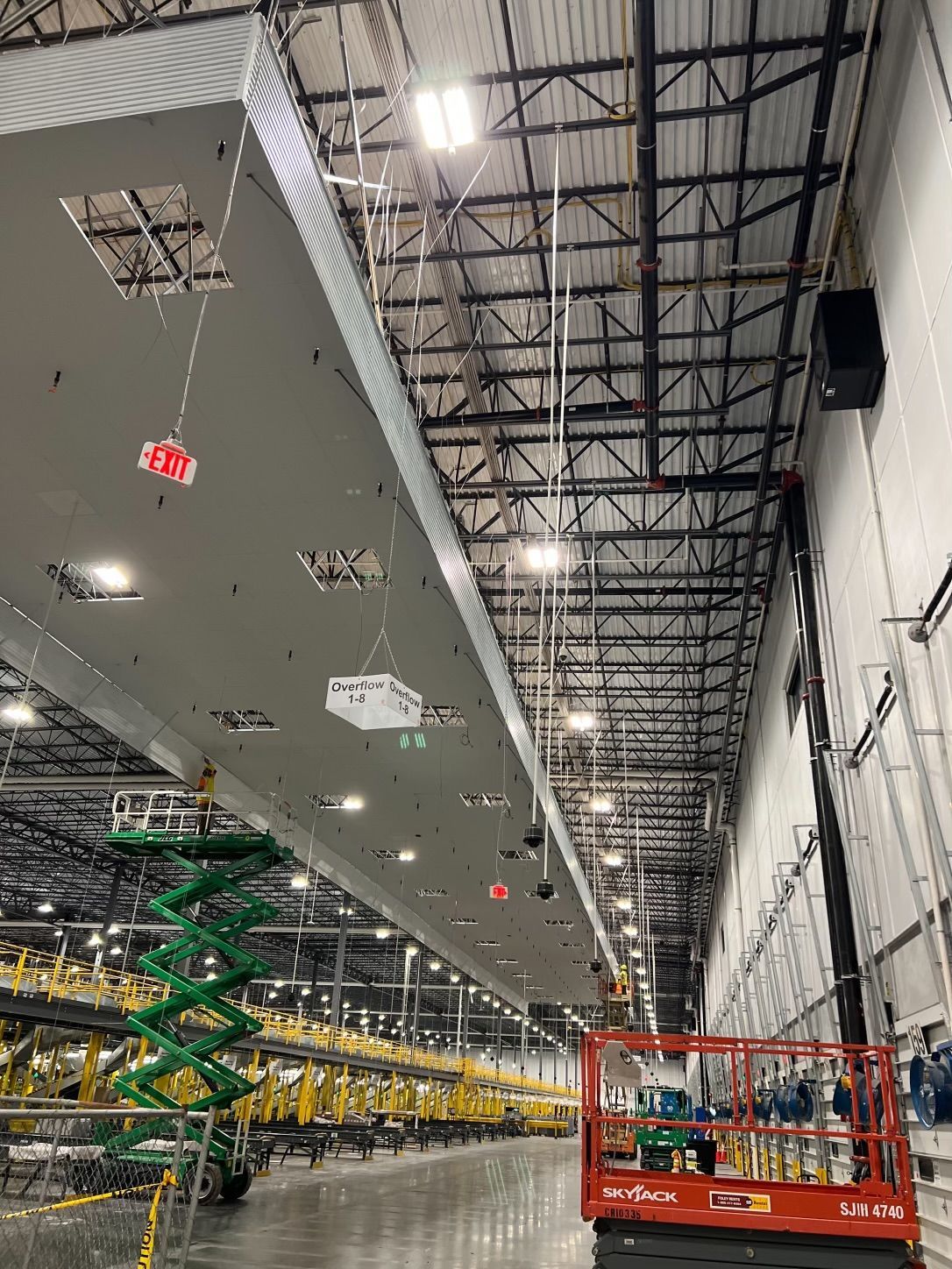 Large industrial warehouse interior with overhead support structures, scissor lifts, and equipment.