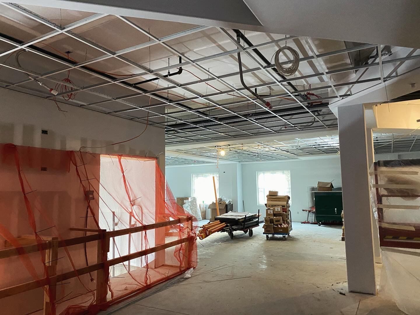 Interior under construction with exposed ceiling grid, orange safety netting, and construction materials.