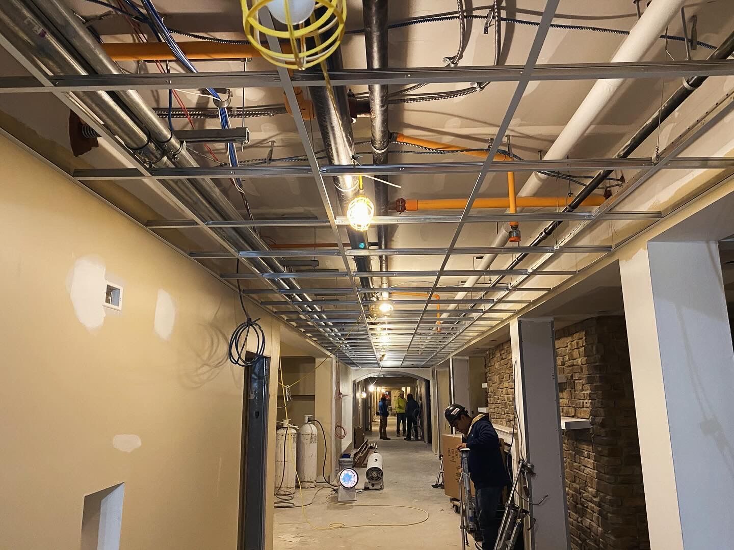 Construction site hallway with exposed ceiling grid, pipes, and lights. Workers and equipment visible.