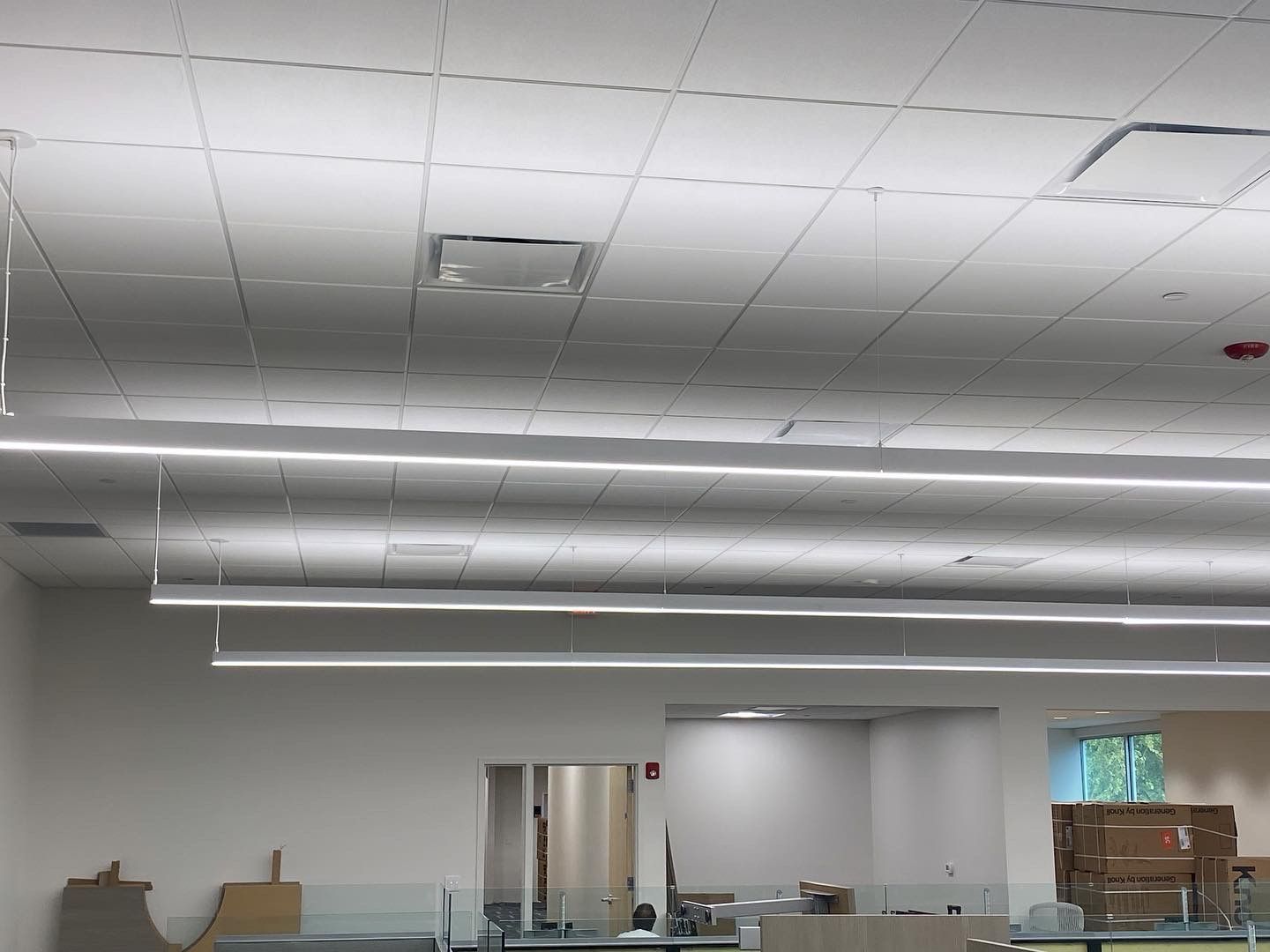 Bright, empty office interior with grid ceiling, suspended lights, and a white wall.