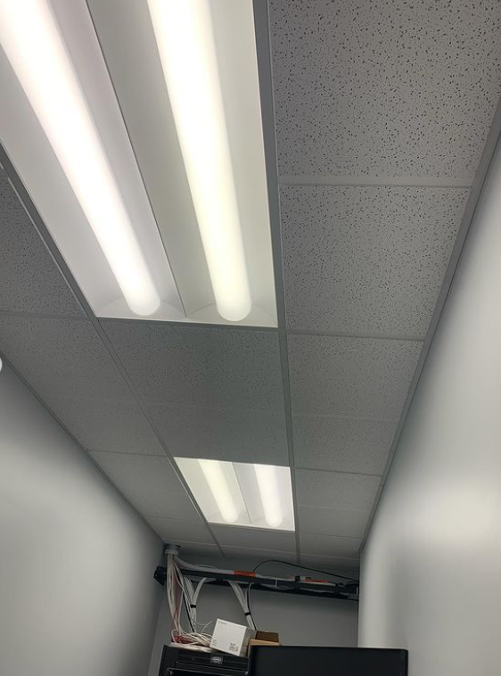 White fluorescent lights in a drop ceiling of a room with white walls, a ladder, and electrical components.