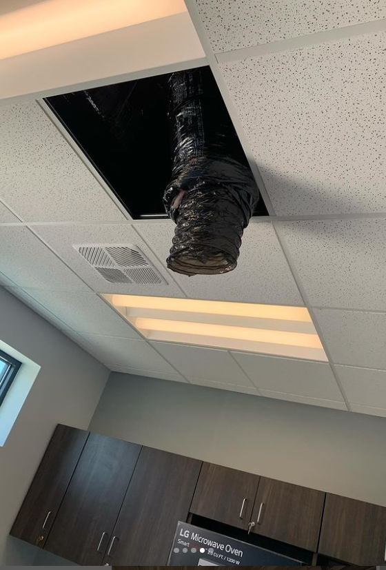 Black vent hanging from an open ceiling panel in a room with brown cabinets.