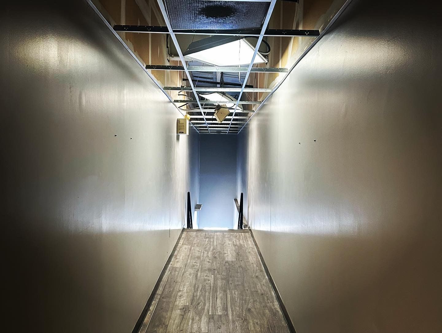 Long, narrow hallway with wooden flooring, walls, and exposed ceiling infrastructure, leading to a doorway at the end.