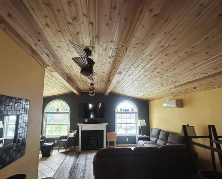 Living room with wood-paneled ceiling, dark grey accent wall, fireplace, windows, and brown sofa.