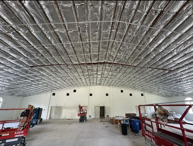 Interior view of a building under construction. Ceiling insulation is installed. Workers and equipment are visible.