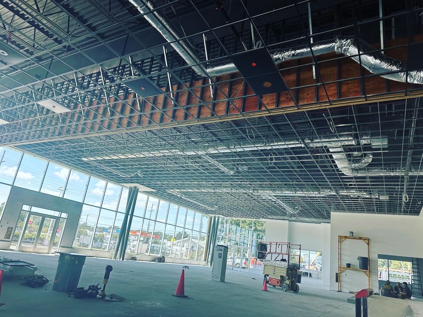 Interior of a building under construction, showing exposed ceiling beams, ductwork, and large windows.