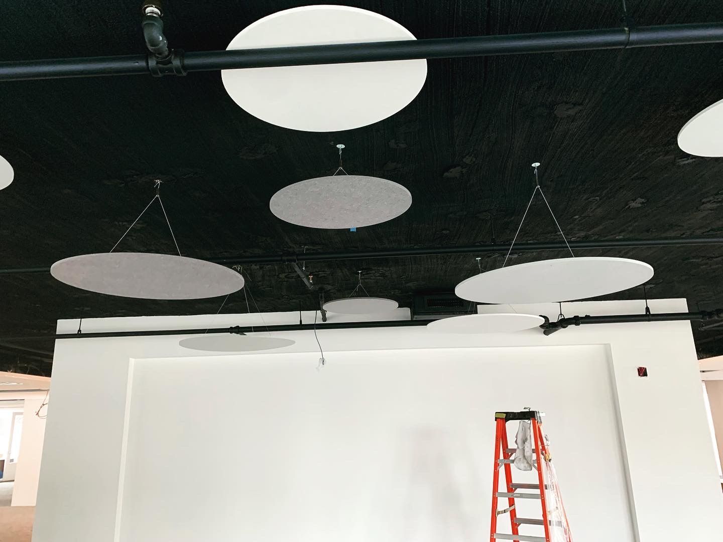 White circular acoustic panels suspended from a black ceiling in a white room; a stepladder is visible.