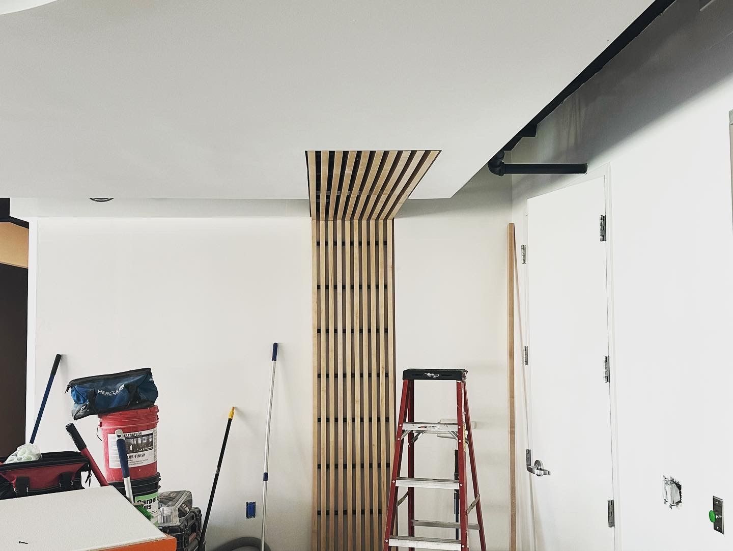 Construction in a room: wood slat wall detail, white walls, ceiling, ladder, tools.