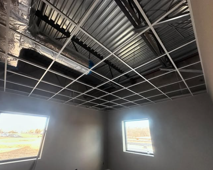 Interior view of a room under construction, with a grid ceiling framework, windows, and exposed ductwork.