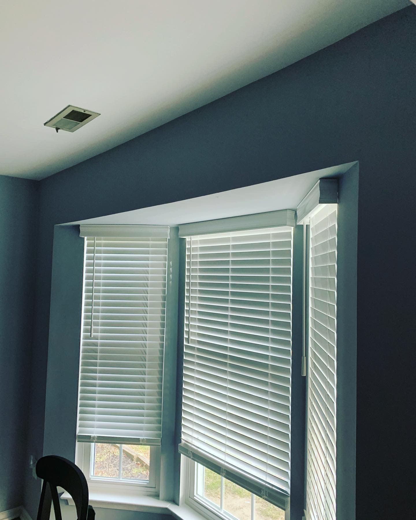 Interior view of a window with white blinds. Blue-gray walls surround the window and a ceiling with a square vent.