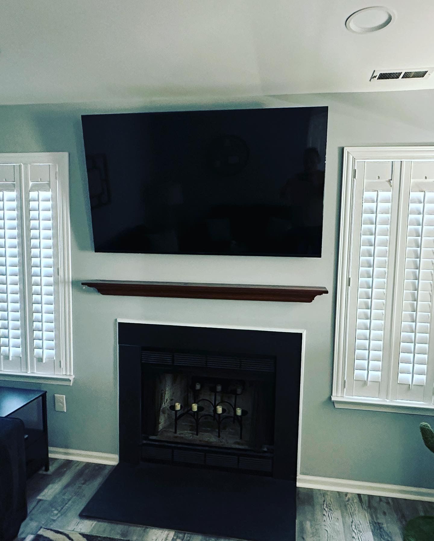 A living room with a fireplace, a mounted TV, and shuttered windows. The fireplace is black with a dark wooden mantel.