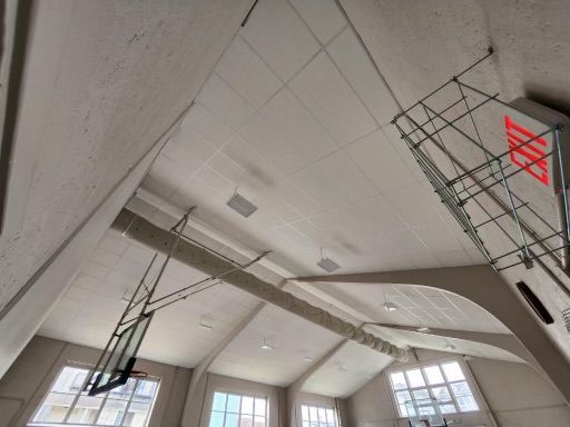 Interior of a gymnasium with basketball hoop and scaffolding against a high, arched ceiling with windows.