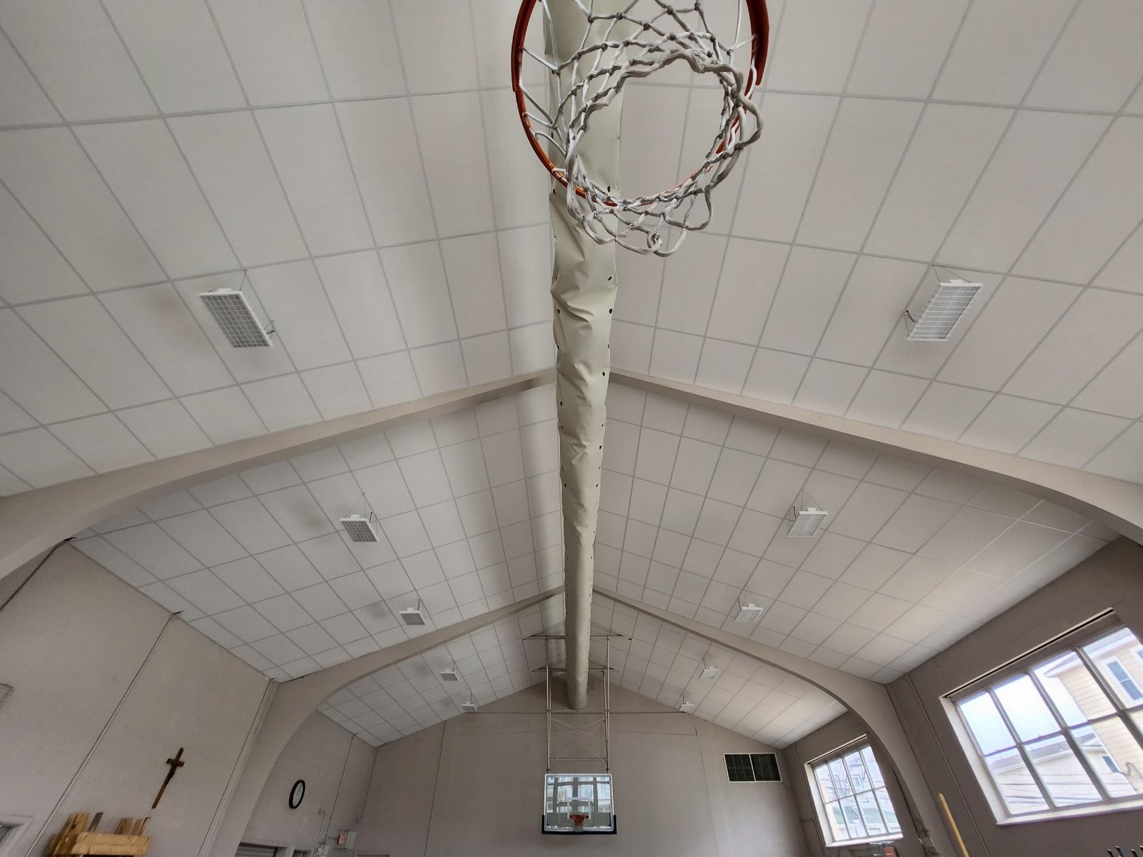 Basketball hoop suspended from a high, vaulted ceiling in a gymnasium with windows.