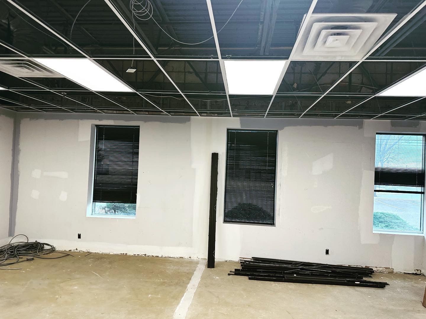 Interior of a room under construction with three windows, drywall, exposed ceiling grid, and concrete floor.
