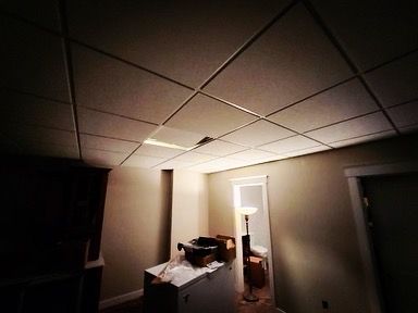 Room with a dropped ceiling, a damaged tile, and a lit doorway. Beige walls and dim lighting.