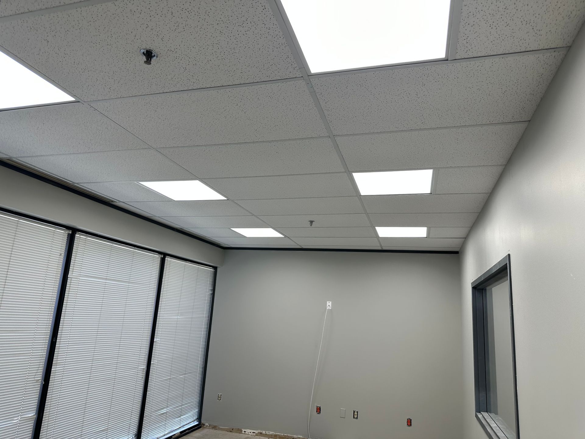 Interior view of an office space with a drop ceiling, fluorescent lights, and large window blinds.
