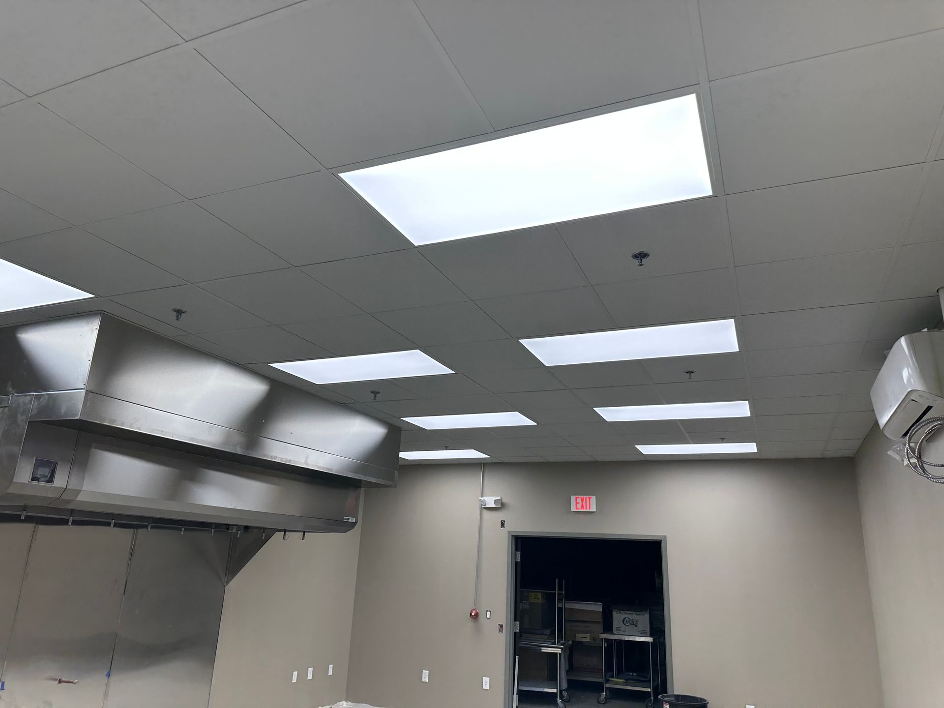 Commercial kitchen interior with a stainless steel vent hood, recessed ceiling lights, and a doorway.