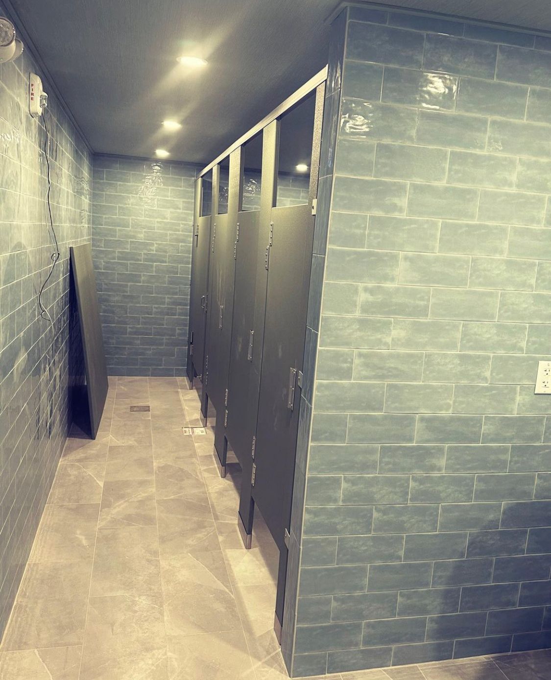 Narrow public restroom with green tiled walls and gray floor, stalls on the right, and overhead lights.