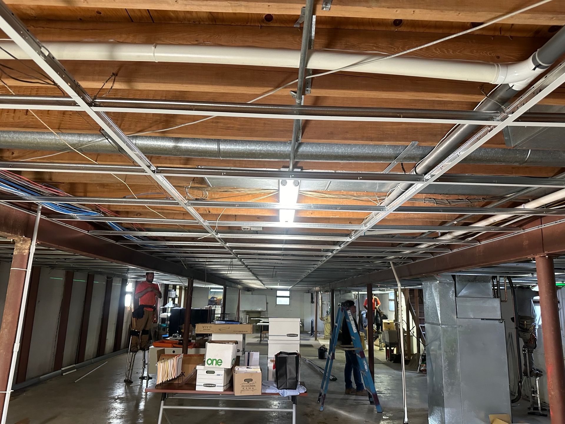 Basement ceiling under construction; metal grid, pipes, and two people working.