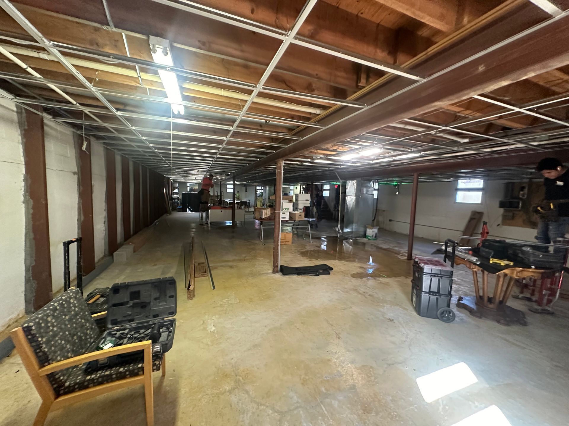 Basement interior with exposed ceiling beams and concrete floor. Tools and chairs scattered about.